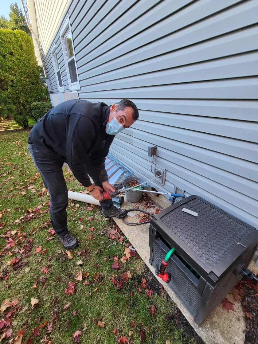 A technician running a rotary brush through an outside dryer vent beside a house